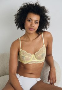 Yellow lace bra with scalloped edges and thin straps, paired with white bikini bottoms featuring lace accents. Smooth, delicate texture.