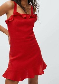 Robe en lin rouge avec encolure à volants et bretelles, corsage ajusté et ourlet évasé. Texture lisse et couleur vibrante.