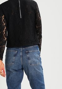 Top lungo in pizzo nero con maniche lunghe, collo rotondo e dettaglio con zip sul retro, abbinato a jeans in denim di un blu chiaro con tasche posteriori.
