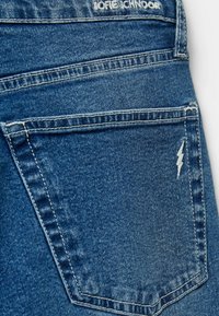 Farkut, joissa on tummansininen pesu, esillä oleva takatasku, jossa on valkoinen salaman muotoinen ommelkoristelu ja näkyvät metalliset yksityiskohdat.