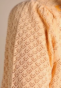 Pull en tricot à motif de dentelle dans un pêche clair, présentant un design texturé avec des formes en diamants et des détails en mailles ouvertes. Tissu doux et fluide.