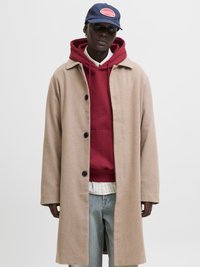 Beige uldfrakke med store sorte knapper, lagdelt over en rød hoodie og en hvid ternet skjorte, parret med lyseblå denim jeans.