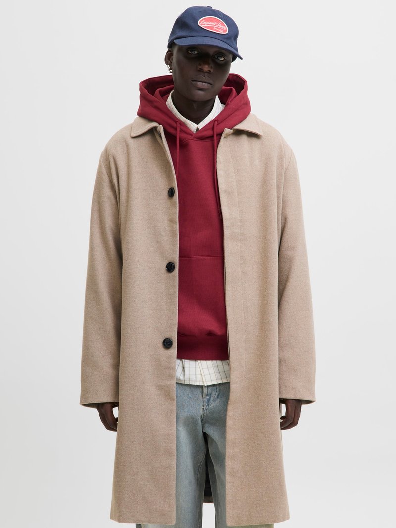 Beige uldfrakke med store sorte knapper, lagdelt over en rød hoodie og en hvid ternet skjorte, parret med lyseblå denim jeans.