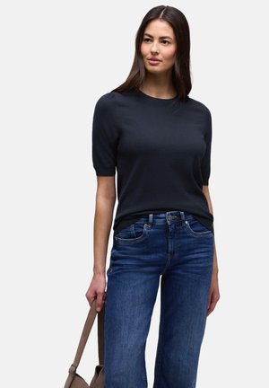 Femme portant un haut noir à manches courtes et un jean bleu, tenant un sac marron à ses côtés, debout devant un fond uni.