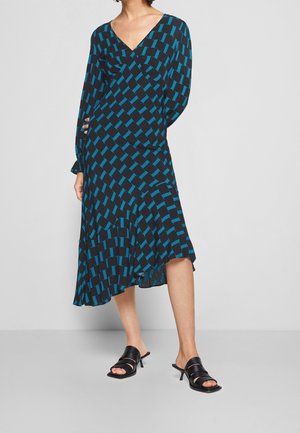 Femme portant une robe midi noire avec un motif géométrique bleu, manches longues, col en V, ourlet asymétrique et sandales à talons noires.