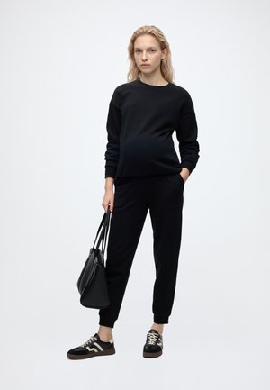 Gravid kvinde iført sort sweatshirt og bukser, der holder en sort håndtaske, står mod en ensfarvet lys baggrund.