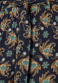 Námořnická tkanina s květinovým paisley vzorem v modrozelené a oranžové barvě. Obsahuje sklady a zavazovací pásek, které ukazují složité detaily a texturu.