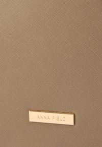 Beežika tekstuuriga pind peene kootud mustriga. Kuldse tooniga kaubamärgi plaat, millele on graveeritud "ANNA FIELD".