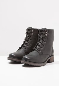 Botas de tobillo de cuero negro con un acabado texturizado, cordones en la parte delantera y cremallera lateral. Presenta un pequeño tacón apilado y suela de goma.