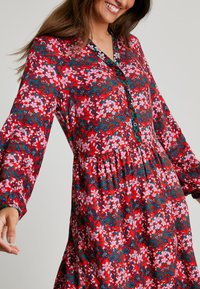 Czerwona sukienka w kwiaty z długimi rękawami, zapięciem na guziki i marszczonym pasem. Ozdobiona różowymi i zielonymi kwiatami na teksturowanej tkaninie.