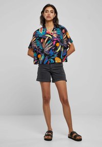Camicia a maniche corte con motivo floreale su sfondo scuro e colori vivaci. Abbinata a pantaloni corti in denim neri e sandali neri.