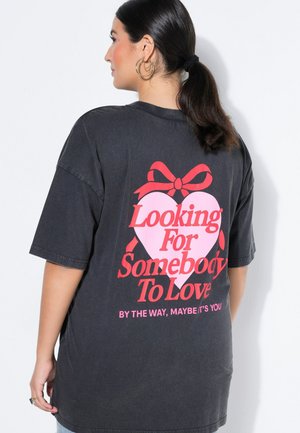 Kobieta z ciemnymi włosami związanymi do tyłu, ubrana w czarny luźny t-shirt z czerwono-różowym wzorem serca i napisem „Looking For Somebody To Love” na plecach.