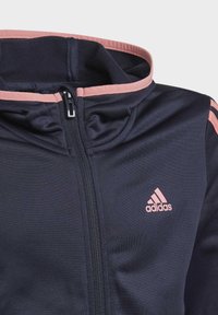 Chaqueta azul marino con cremallera y un acento rosa en el cuello y el hombro. Presenta un logo en el pecho y una tela lisa y texturizada.