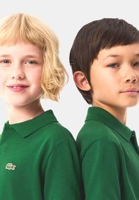 Dwoje dzieci ubranych w pasujące zielone polo z teksturowanej tkaniny. Każda koszulka ma kołnierzyk i logo Lacoste po lewej stronie.