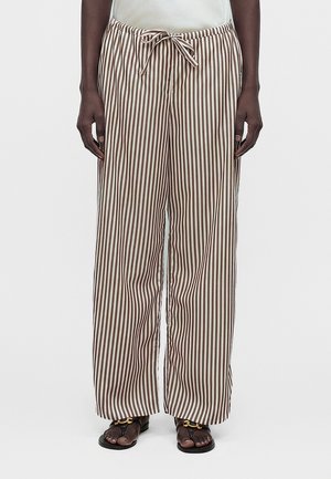 Άτομο που φοράει φαρδιά, μπεζ και καφέ κάθετα ριγέ παντελόνια με κορδόνι στη μέση και μαύρα σανδάλια ανοιχτά στη μύτη με χρυσή λεπτομέρεια.