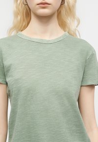 Personne blonde portant des boucles d'oreilles créoles argentées et un t-shirt vert clair à manches courtes et col rond sur un fond blanc.