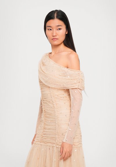 Vrouw met lang, donker haar in een beige, geplooide off-shoulder jurk versierd met sequins, staand tegen een eenvoudige witte achtergrond.