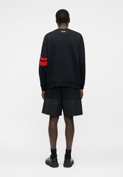 Άνδρας με μαύρο φούτερ με κόκκινες ρίγες στο αριστερό μανίκι και μαύρο σορτς, στέκεται με την πλάτη προς τα εμάς, μπροστά σε ένα απλό φόντο.