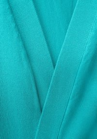 Turkos stickad tyg med en ribbad struktur, med överlappande tygsektioner som skapar en V-form. Slät och lätt utseende.