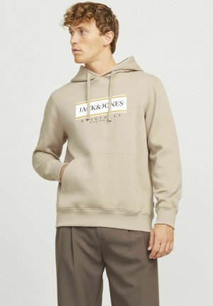 Haut à capuche beige en tissu doux, avec une poche frontale, une capuche à cordon et un logo imprimé en couleurs contrastées sur la poitrine.