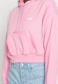 Růžová oříznutá mikina s kapucí z měkké látky, vybavená přední kapsou, stahovacími šňůrkami a bílým logem "Levi's". Kombinována s světle modrými džínami.