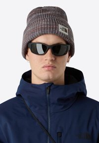 Persona che indossa un berretto lavorato a maglia grigio con un logo, occhiali da sole neri, orecchini a cerchio in argento e una giacca zip navy con colletto alto.