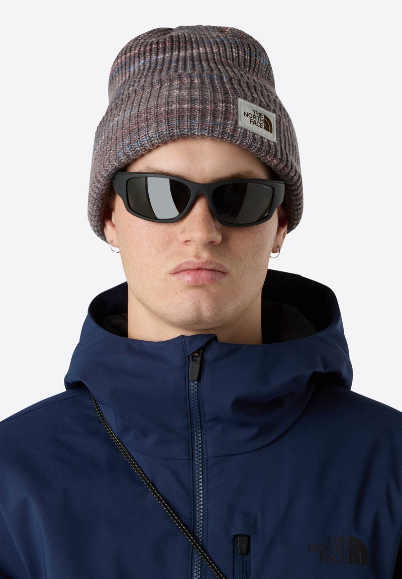 Persona que lleva un gorro de punto gris con un logo, gafas de sol negras, aretes de aros plateados y una chaqueta azul marino con cremallera y cuello alto.