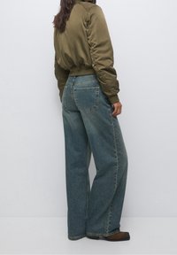 Mujer vestida con una chaqueta bomber verde oliva, jeans de pierna ancha azules y botas marrones, de pie contra una pared gris, mirando hacia afuera.