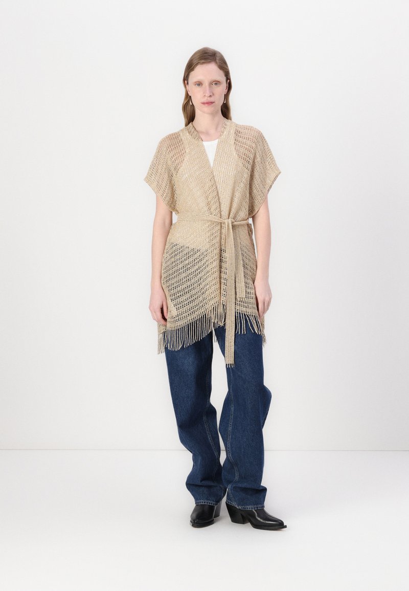 Beige neulecardigan, jossa on lyhyet hihat, hulmuava helma ja solmittava vyö. Päällä valkoinen toppi ja parina leveälahkeiset farkut.
