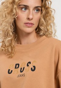 Beżowy sweter z ribbowanym dekoltem, z napisem "CROSS" w kolorze czarnym i akcentami z kryształków, oraz z mniejszym napisem "JEANS" poniżej, również w kolorze czarnym.