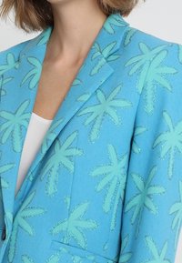 Kobieta ubrana w jaskrawoniebieską marynarkę z jasnoniebieskimi wzorami palm oraz białą bluzkę pod spodem, przyciętą na wysokości szyi i ramion.