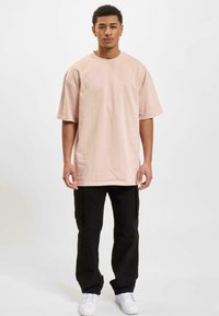 Lys rosa oversized t-shirt med korte ærmer og rund halsudskæring, parret med sorte cargo-bukser og hvide sneakers på en model foran en ensfarvet baggrund.
