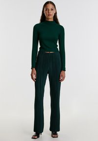 Donkergroene geribde langemouwtop met gegolfde halslijn, gecombineerd met geplooide wijde broek in dezelfde kleur, gedragen met zwarte open teen hakken.