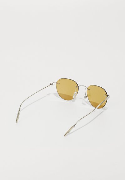 Lunettes de soleil dorées avec monture en métal, verres géométriques et branches fines. Les caractéristiques notables incluent des plaquettes de nez réglables et un design minimaliste.