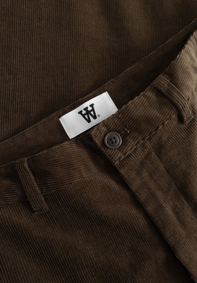 Pantalons en velours côtelé marron avec une surface texturée, présentant une fermeture à boutons et une étiquette blanche avec un logo noir.