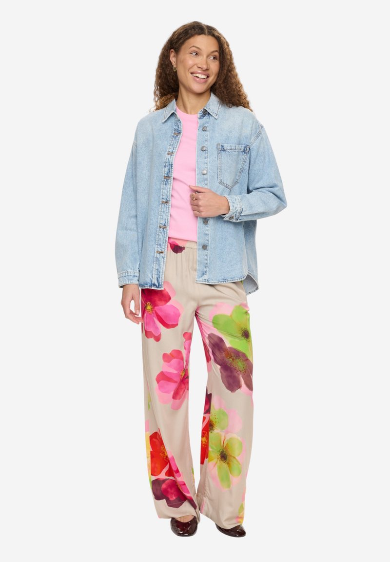 Femme souriante portant une veste en denim bleu clair, une chemise rose, un pantalon large beige avec de grandes impressions florales colorées, et des chaussures sombres.