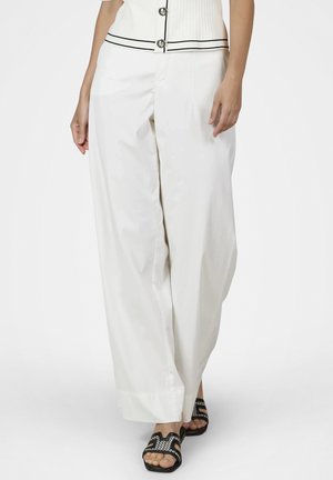 Woman wearing white wide-leg pants and black open-toe sandals with woven detailing, standing against a plain white background.