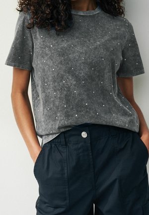 Personne portant une chemise grise à manches courtes avec de petits points blancs et un pantalon bleu foncé boutonné, les mains dans les poches.