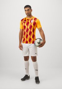 Maglietta da calcio con un vivace motivo a onde arancioni e rosse, abbinata a pantaloni corti bianchi, scarpe da calcio nere e calze bianche fino al ginocchio. Tiene un pallone da calcio.