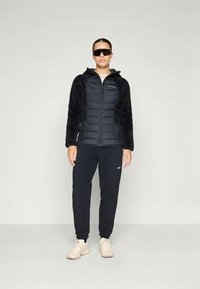 Čierna prešívaná bunda s umelými kožušinovými rukávmi, so zipsom a stojatým golierom, skombinovaná s čiernymi nohavicami a svetlými topánkami. Model nosí slnečné okuliare.