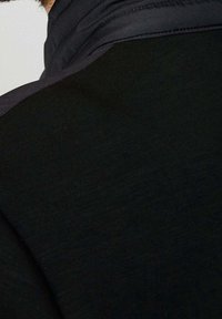 Personne portant une chemise noire à manches longues avec une veste sombre posée sur une épaule, cadrée de près sur le haut du dos et la nuque.