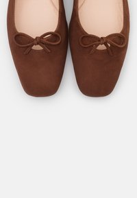 Ballerines en daim marron avec un bout arrondi, présentant un petit nœud à l'avant et une doublure intérieure lisse.