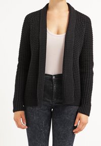 Schwarze, gewebte Strickjacke mit einem strukturierten Muster, offener Vorderseite und gerippten Ärmelabschlüssen. Kombiniert mit einem hellen Oberteil und dunklen Jeans.