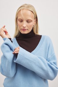 Hellblaues, übergroßes Sweatshirt mit gerippten Bündchen und V-Ausschnitt. Darunter wird ein dunkelbrauner Rollkragenpullover getragen. Weicher Stoff, lockerer Sitz.