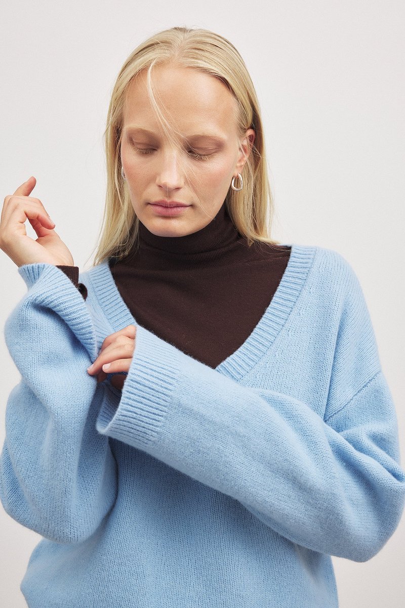 Hellblaues, übergroßes Sweatshirt mit gerippten Bündchen und V-Ausschnitt. Darunter wird ein dunkelbrauner Rollkragenpullover getragen. Weicher Stoff, lockerer Sitz.
