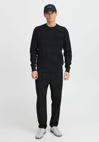 Marineblå strikkegenser med en teksturert, bølgete mønster, kombinert med svarte bukser og grå sneakers. Svart caps med hvite tekstdetaljer.