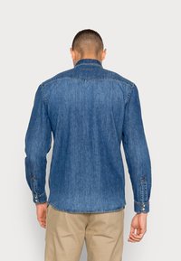 Man met zijn rug naar de camera, gekleed in een langwerpig blauw spijkerhemd en beige broek, tegen een effen lichte achtergrond.