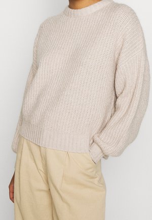 Personne portant un pull en maille texturé couleur crème à manches longues et un pantalon beige taille haute, main dans la poche.
