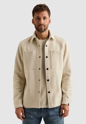 Homme portant une veste beige boutonnée sur un pull léger en maille et un jean bleu, debout devant un fond clair uni.