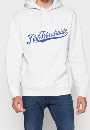 Jasnoszara bluza z kapturem z niebieskim napisem "Huf Worldwide" na piersi, noszona z ciemnoniebieskimi dżinsami przez stojącą osobę z luźno opuszczonymi rękami.